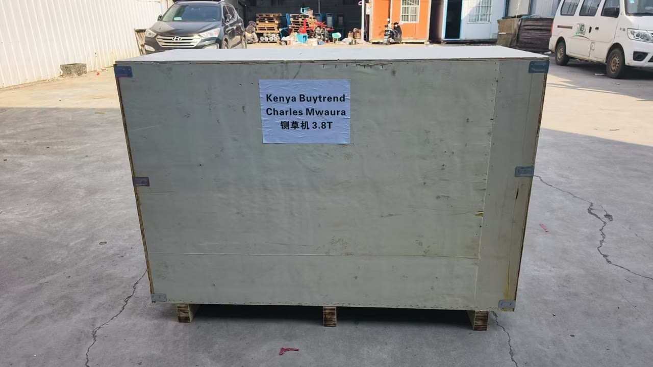 A pale plywood export crate standing in a factory yard, labelled Charles Mwaura, chaff cutter 3.8T, for Kenya, with vehicles and the workshop behind it